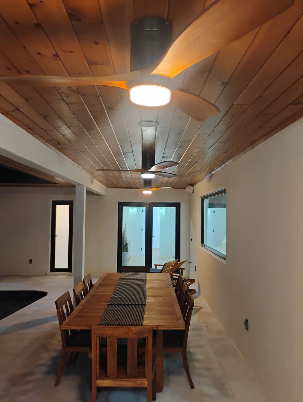 Modern dining area with wooden table, ceiling fans, and large windows. Minimalist design.